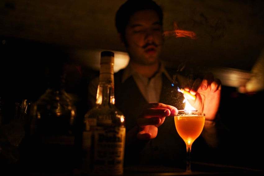 Jody Horton's image of a man with a theatrical mustache preparing a cocktail in a dimly lit room. He holds a lit match over the cocktail glass.