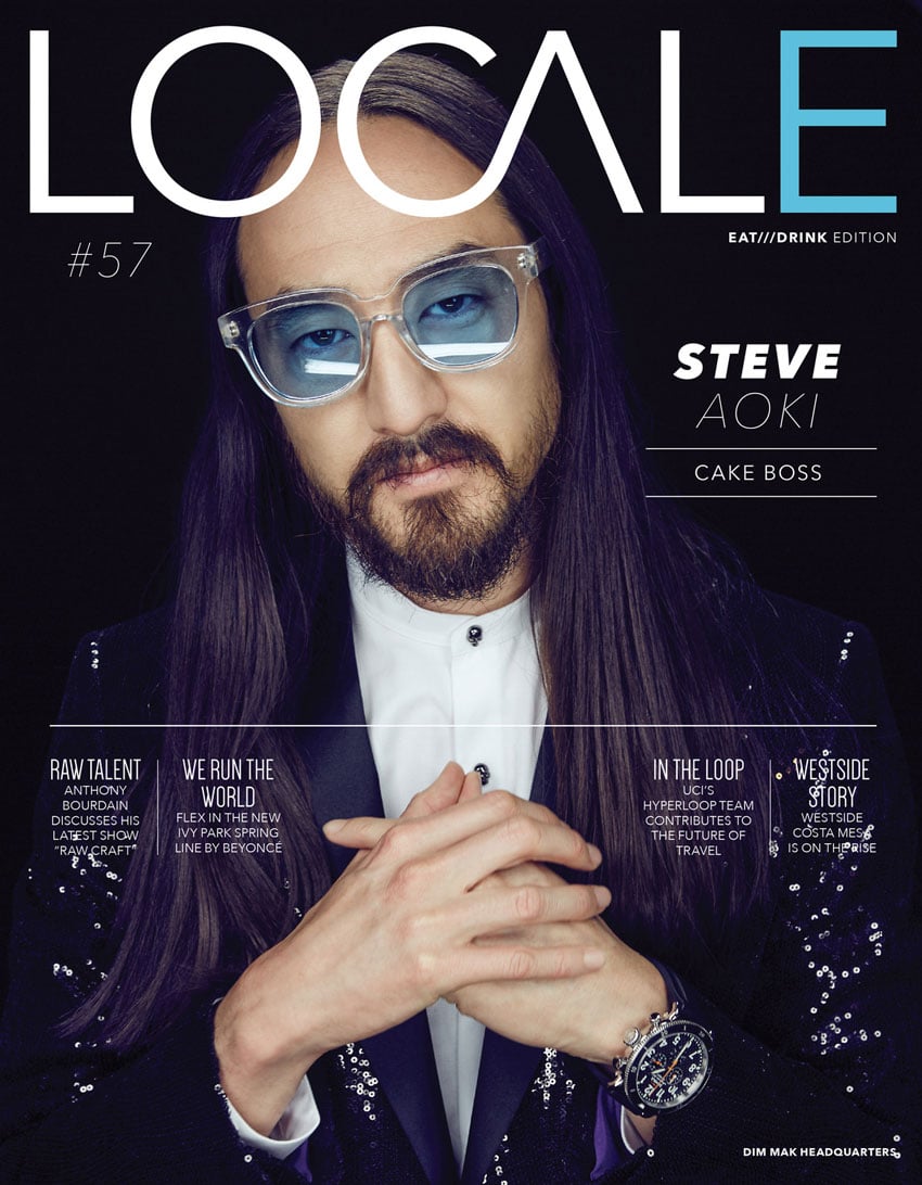 A photo by JSquared for the cover of Locale Magazine featuring a portrait of Steve Aoki. Steve looks directly at the camera with a neutral expression. He has long dark hair and wears glasses with bold clear frames with blue tinted lenses. His hands are folded together in front of his chest. 