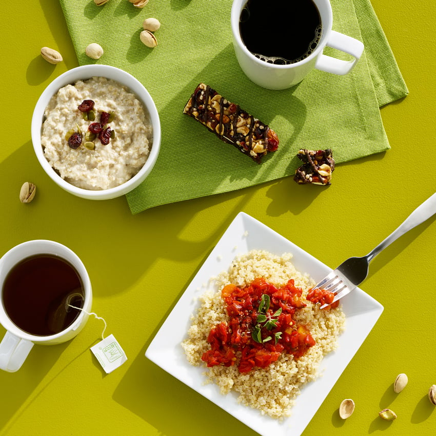 Kyle Dreier's photograph for Lifebox of a flat lay containing a couscous dish, cups of tea and coffee, a snack bar, a small bowl of oatmeal, and a handful of pistachios on a chartreuse surface.