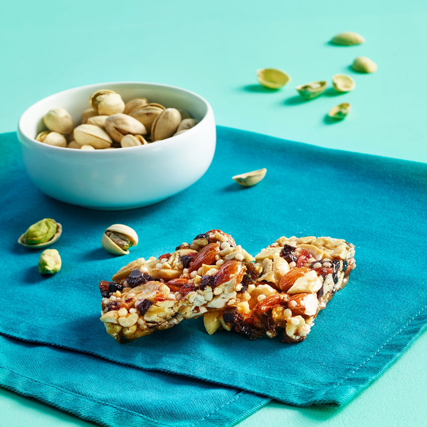 Kyle Dreier's photograph for Lifebox of a small bowl of pistachios and a fruit and nut snack bar broken in half on a teal cloth napkin on a robin's egg blue surface. Some pistachios and pistachio shells are strewn about the napkin and surface.