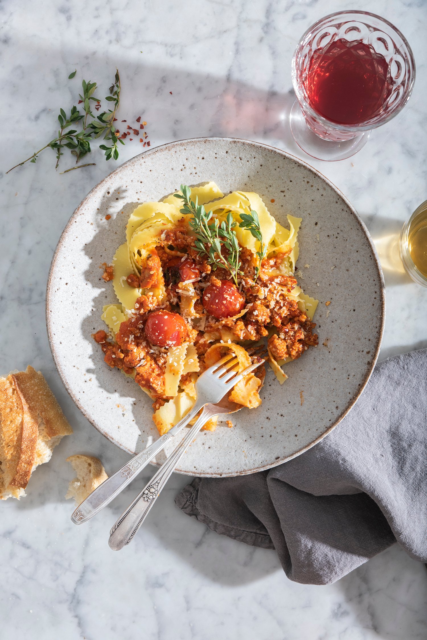 Pasta noodles with meat sauce and herb garnish plated on rustic large grey and earth-tone bowl with brown rim accompanied by a glass of white wine and torn piece of baguette 