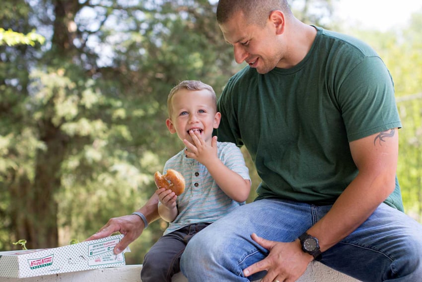 Photographer Liz Nemeth's photo for Krispy Kreme's social media campaign. The photo features a man and a young boy sitting together outside. The little boy smiles with a glazed donut, and the man smiles at the boy while reaching around his back to touch a Krispy Kreme box.