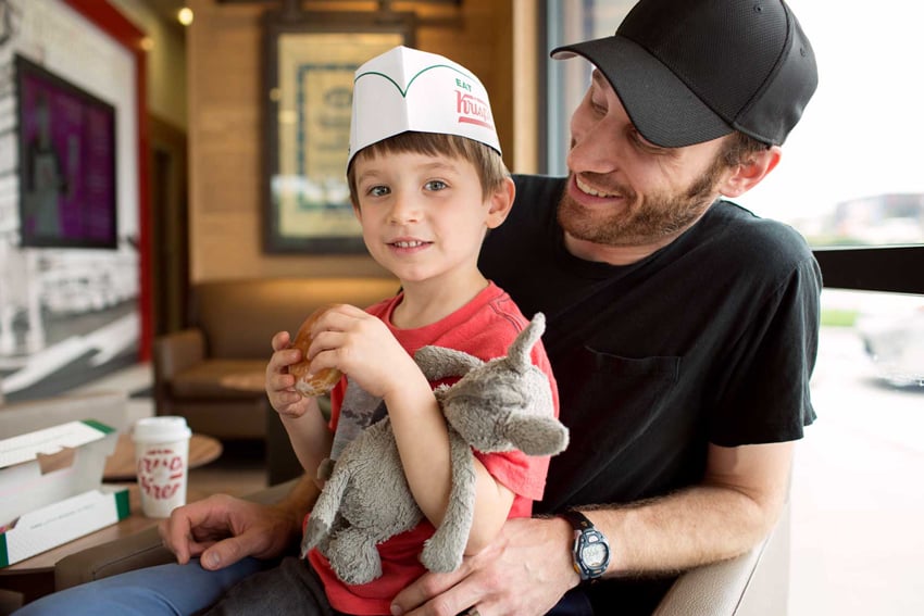 Photographer Liz Nemeth's photo for Krispy Kreme's social media campaign. The photo features a man wearing a black t-shirt and baseball cap with a young boy on his lap. The boy wears a paper Krispy Kreme hat and holds a tattered stuffed animal in the crook of his elbow. He holds a donut and smiles directly at the camera. There is an open Krispy Kreme box in the background and a to-go cup for a hot beverage.