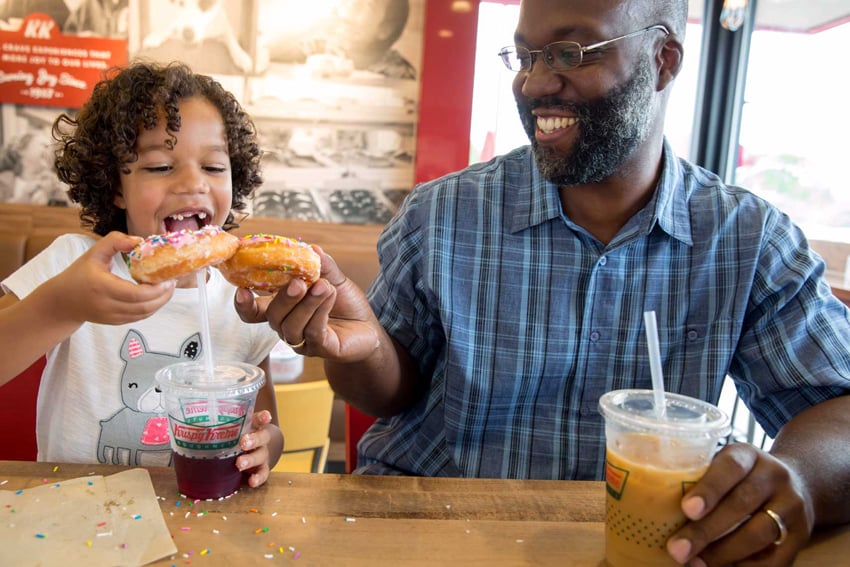 Photographer Liz Nemeth's photo for Krispy Kreme's social media campaign. The photo features a man and a young girl sitting in a Krispy Kreme shop and smiling. They each have their left hands on cold drinks in Krispy Kreme clear plastic cups and use their right hands to cheers their donuts that have pink frosting and colorful sprinkles.