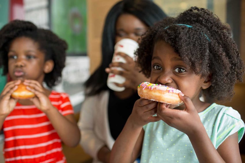 Photographer Liz Nemeth's photo for Krispy Kreme's social media campaign. The photo features three people: two children and one woman. One child is in focus in the foreground eating a donut with pink frosting and colorful sprinkles. She wears a light blue top.