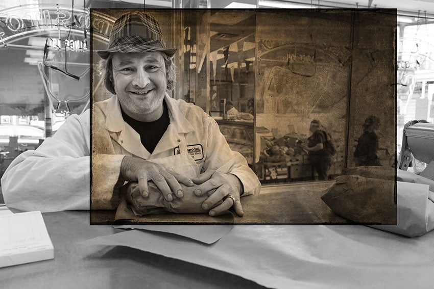 Nashville-based photographer Mark Boughton's photo from Philadelphia's Italian Market. The black and white portrait features a man wearing a plaid fedora and a white work jacket and apron, smiling warmly from behind a deli counter. Between his hands on the counter is a parcel wrapped in paper.