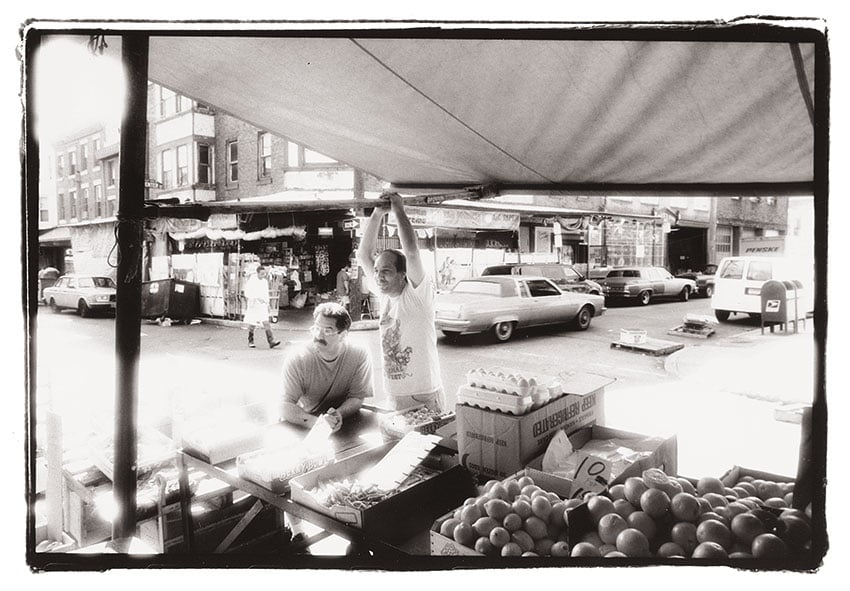 Nashville-based photographer Mark Boughton's photo from Philadelphia's Italian Market. The black and white photo features two men in relaxed positions under an awning in the market. Also under the awning are boxes of produce and cartons of eggs.