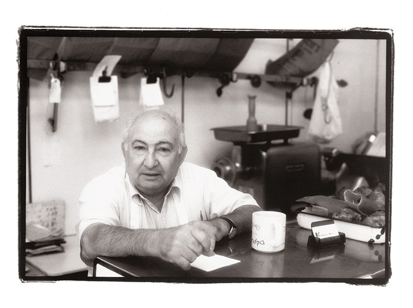 Nashville-based photographer Mark Boughton's photo from Philadelphia's Italian Market. The black and white portrait features an elderly man behind a deli counter. He has a neutral facial expression, dark eyebrows, and white hair, and he looks directly at the camera. 