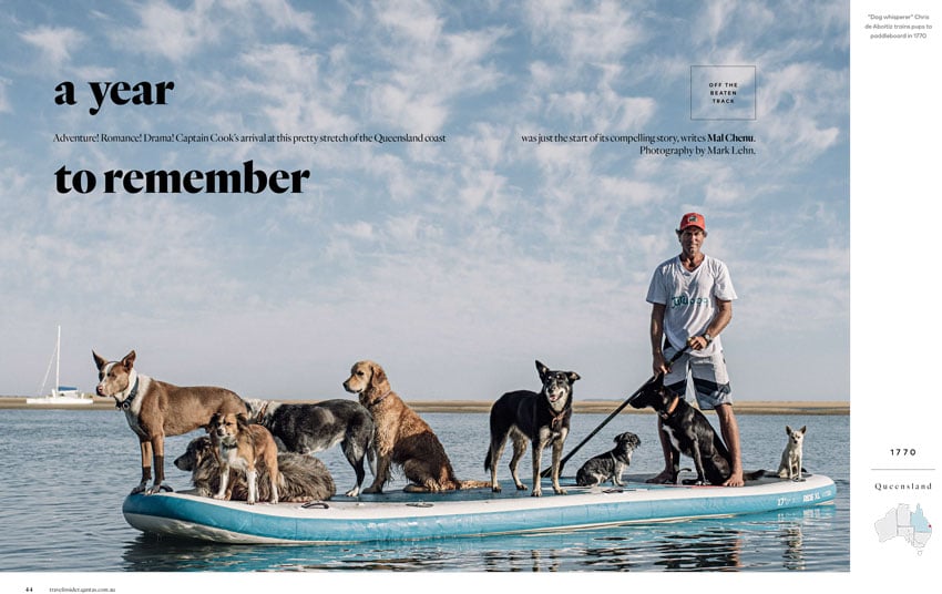 A tear sheet for Qantas Travel Insider by photographer Mark Lehn of a man in an orange baseball cap holding a paddle on a paddle board with 9 dogs. The waters are calm, and there is a sail boat in the background.