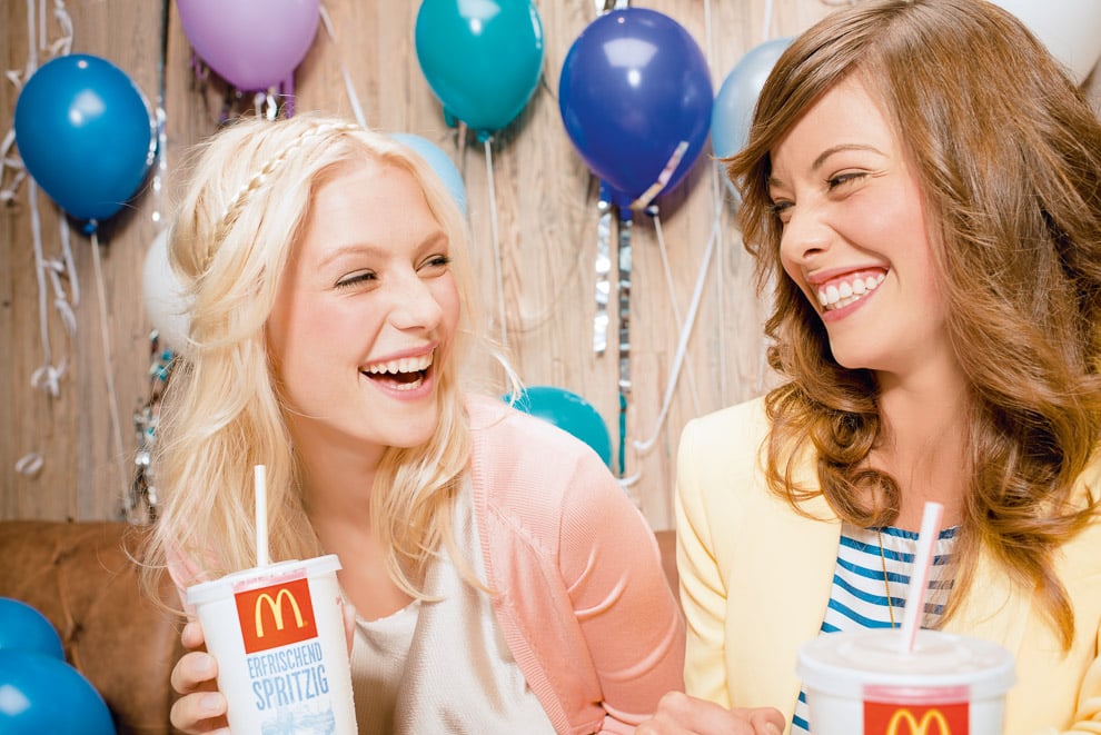Two friends smiling at each other while sipping McDonald's sodas; balloons and streamers behind them shot by Munich-based lifestyle photographer Christian Brecheis 