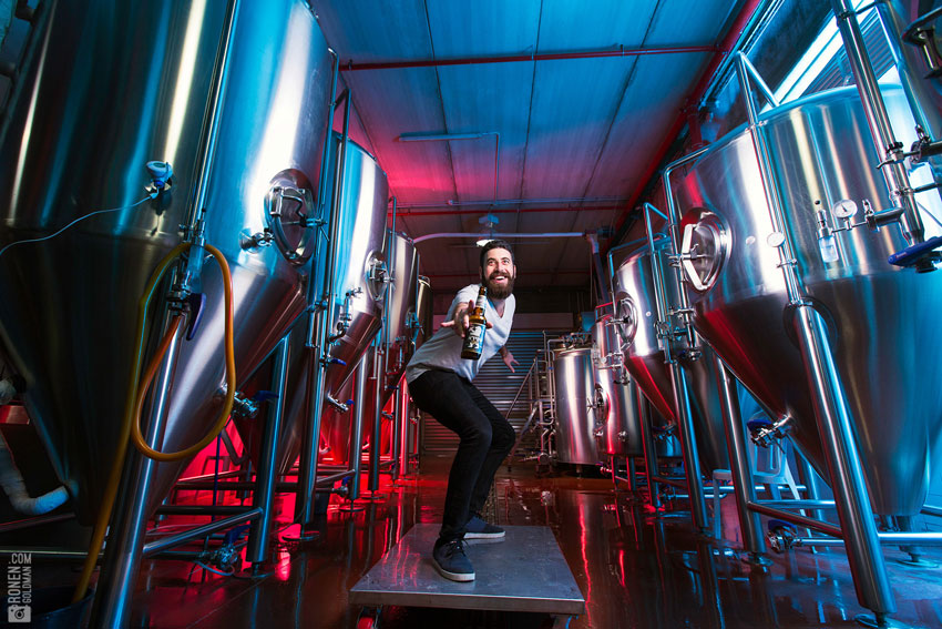 A man posing with a bottle of Shapiro beer in a brewery surrounded by fermentation tanks, photo by Ronen Goldman.