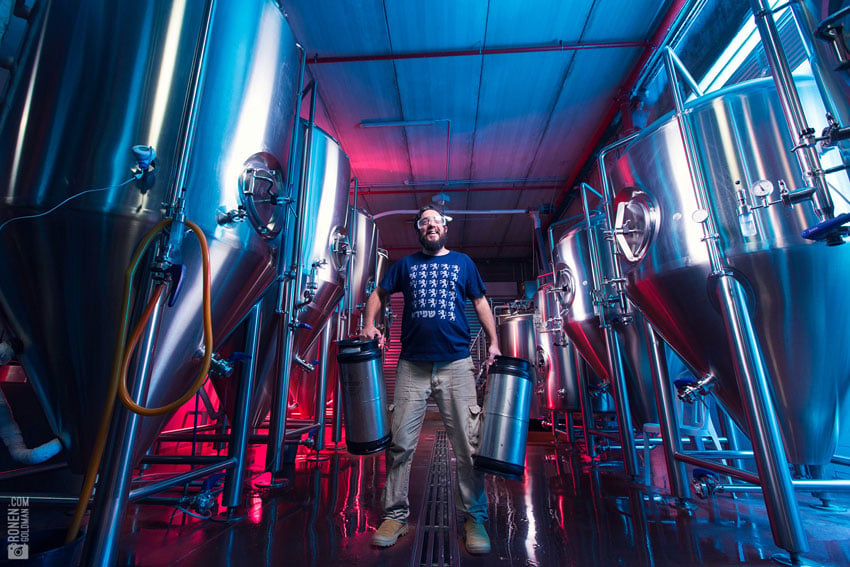A man posing in a brewery surrounded by fermentation tanks, photo by Ronen Goldman.