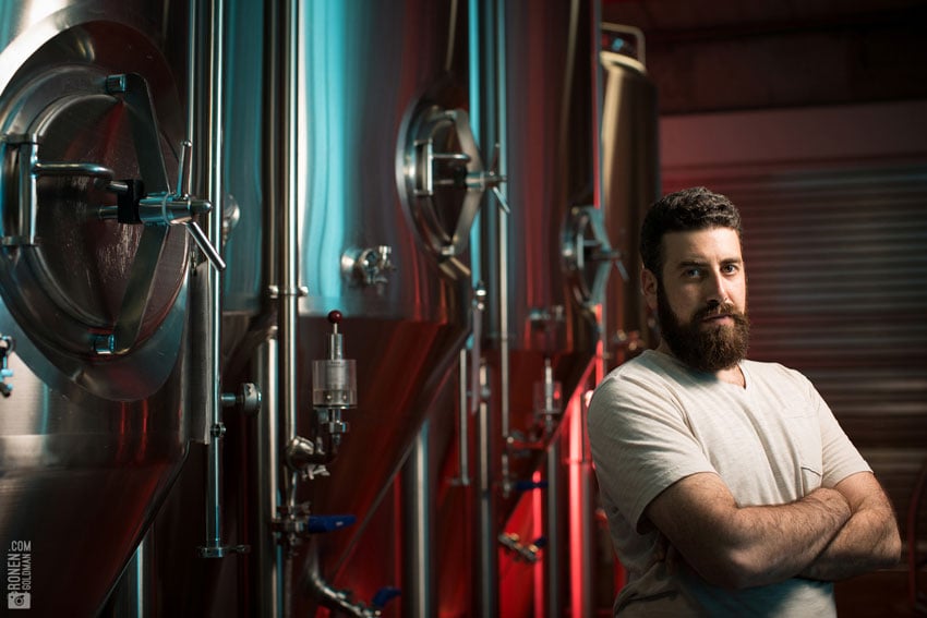 A portrait of a man in a brewery surrounded by fermentation tanks, photo by Ronen Goldman.
