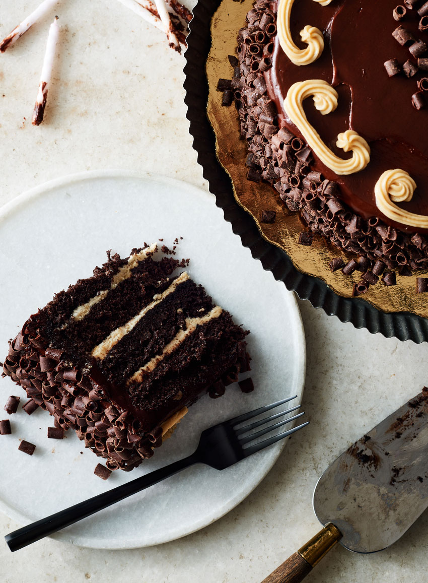 An image by photographer Stephen DeVries for Publix's Gather and Share magazine. The photo, taken from above, features a chocolate cake in the upper right corner. It is partially out of the frame. The cake is round, and garnished around the sides with chocolate shavings and around the top edges with white icing designs. On the left, slightly out of frame is a white plate holding a slice of the cake on its side and a metal fork. In the lower right corner is a cake knife with chocolate smudges on it, and in the upper left corner are a handful of white partially burnt candles with chocolate smudges at their bases.