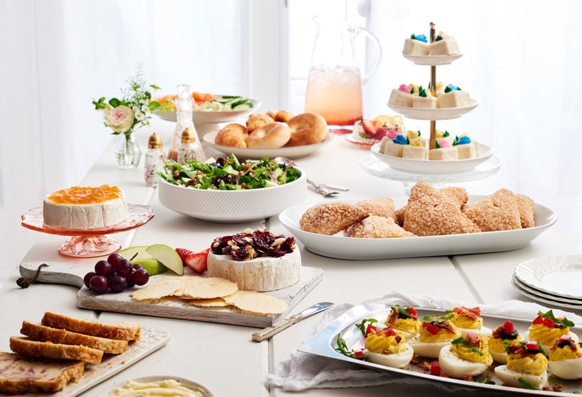 An image by photographer Stephen DeVries for Publix's Gather and Share magazine. The photo features several dishes atop a white table with a brightly lit white background of white windows, white walls, and transparent white curtains. The dishes include a platter of deviled eggs, a plate of bagels and croissants, a pitcher filled with a light pink beverage and ice, a bowl of salad, and a three-tiered serving piece of petite-fours. There are also two different wheels of soft cheese.