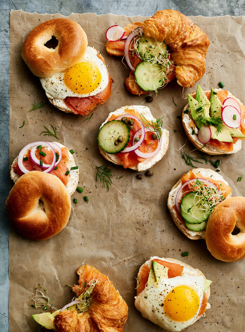 A colorful photo taken from above by photographer Stephen DeVries for Publix's Gather and Share magazine. The image features several bagel and croissant sandwiches and open-faced sandwiches with ingredients like cream cheese, salmon, capers, slices of red onion, cucumbers, microgreens, fried eggs, herbs, and cracked pepper. The food is laid out on a piece of parchment paper laid over a metal surface.