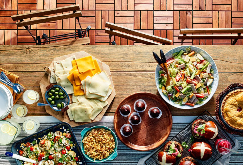 A colorful photo by photographer Stephen DeVries for Publix's Gather and Share magazine. The photo is taken from above and features various dishes laid out on a wooden table with wood metal folding chairs along its side. The dishes include a platter of cheese slices with a small bowl of olives, a salad, a pie, a roasted vegetable dish, a platter with some sort of chocolate dessert, and a rack with some small sandwiches on pretzel buns. There is also a jug and three glasses filled with lemonade.
