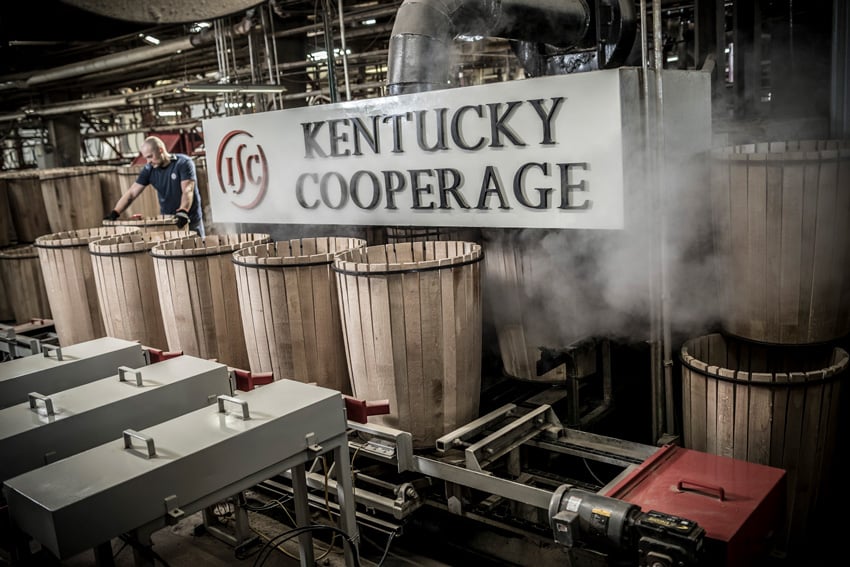 A photo taken by photographer Tadd Myers for The Independent Stave Company. In the center of the photo is a large white sign that says "Kentucky Cooperage" in black with the company's red "ISC" logo. The sign is situated amongst metal factory machinery and a number of wooden barrels that are in the process of being made. There is steam in the air. To the left of the sign is a man wearing a t-shirt and working gloves handling one of the barrels.