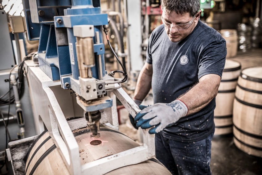 A photo by Tadd Myers of a worker at ISC using machinery to bore a hole into a wooden barrel. The worker is a man wearing a navy blue t-shirt with a small white ISC logo on the left side of the chest and denim pants. He wears protective eyewear and protective gloves.