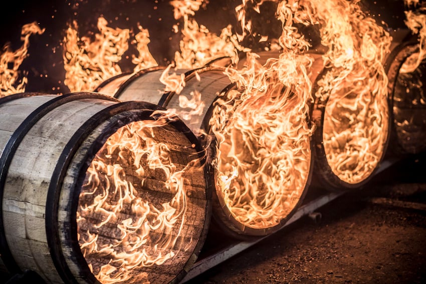 A photograph by Tadd Myers of a row of wooden barrels being flame charred. The barrels are positioned on their sides on a sort of rack, and the flames are concentrated inside the barrels.