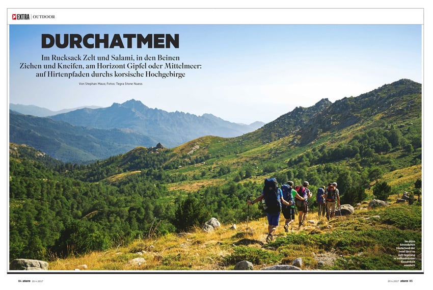 A tear sheet by Photographer Tegra Stone Nuess for Stern Magazine. The photo features five people with hiking backpacks and hiking poles trekking in single file along a grassy, stoney path with an expansive green mountainous background with blue skies.