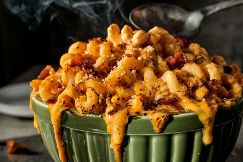 Photographer Teri Campbell's image of a bowl of macaroni and cheese with pieces of bacon and bread crumbs in a ridged green ceramic bowl. A spoon is poised over the bowl and steam rises from the food.