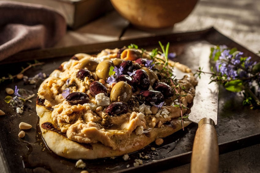 A photo taken by photographer Teri Campbell of a piece of pita bread spread with hummus. There are brown and green olives on top of it as well as some crumbles of white cheese and sprigs of herbs. The pita sits on top of an oiled cookie sheet, and there are tiny purple flowers sprinkled on and around the pita.