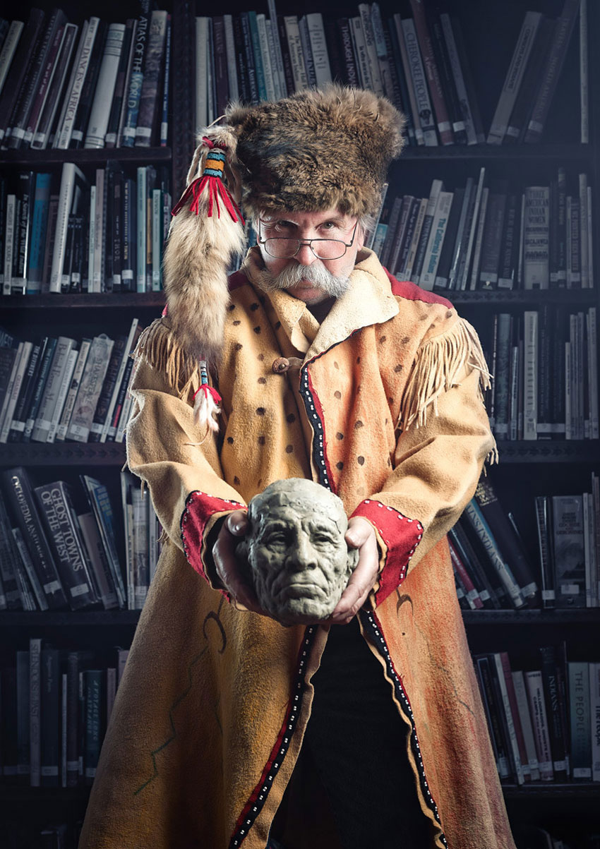 Photographer Willie Petersen's photo for his collaboration entitled "Inspiration to Creation: The Hidden Life Behind Bronze." The photo features a white man wearing aviator style glasses and a Native American style buckskin jacket and raccoon skin hat. The man looks directly at the camera and holds out a wax model of an elderly man's head in both hands in front of him.