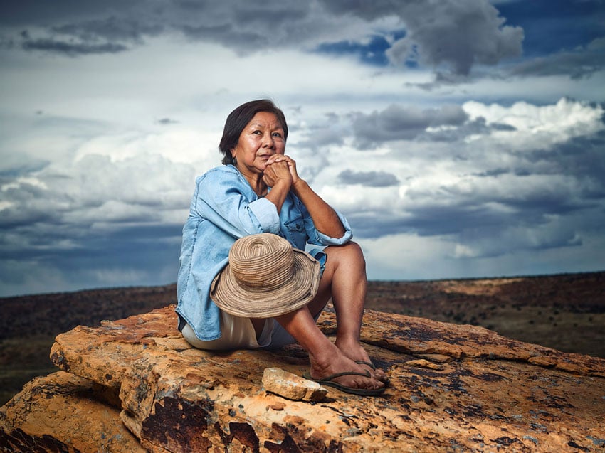 Willie Petersen's photo of a woman in a blue chambray button down top, khaki shorts, and flip flops. She has brown skin and short black hair. She sits atop a natural stone in front of an expansive arid Arizona landscape. Her knees her elbows rest atop her knees which are pulled up in front of her, and her hands are clasped together under her chin as she looks into the distance over the photographer.