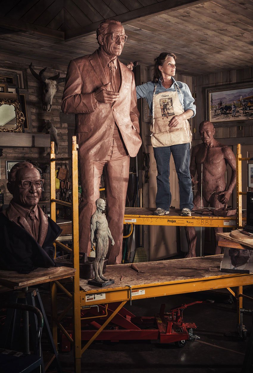 Willie Petersen's portrait of a woman wearing a denim outfit and a work apron standing on a yellow scaffold among a number of sculptures. Her right hand rests on the left shoulder of a larger than life sculpture of a man  and she looks off into the distance to her left.