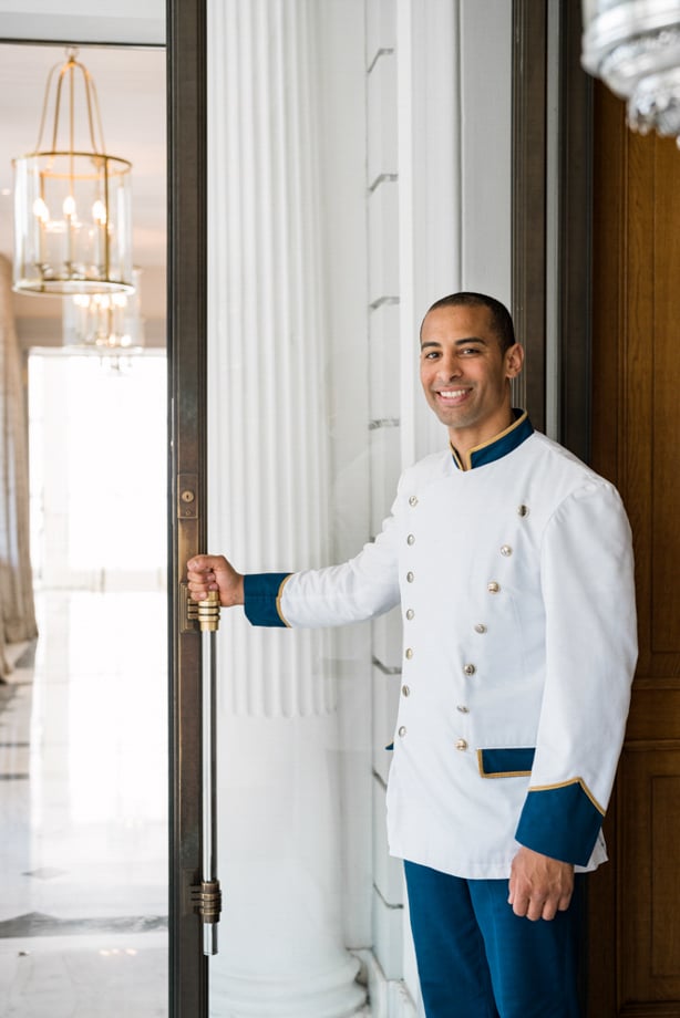 Creative in Place: Be My Guest photographer Clara Tuma's image of a smiling bellhop holding a glass door's handle. He wears a double breasted white jacket with gold buttons and dark teal and gold details with dark teal slacks.