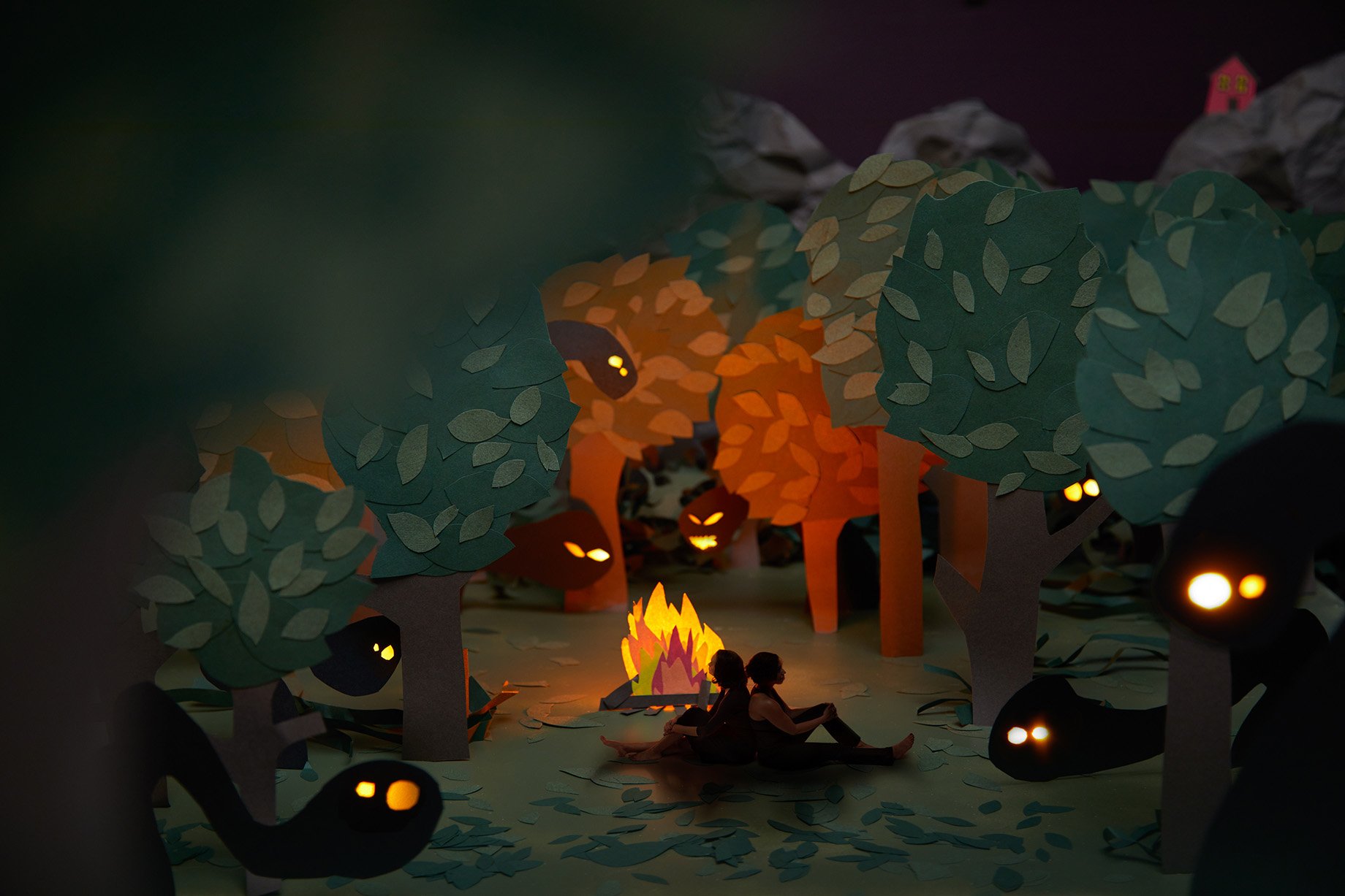 Two woman sit back to back at a campfire in the woods while spooky creatures watch made from construction paper shot by Patrick Heageny for Paper Thin