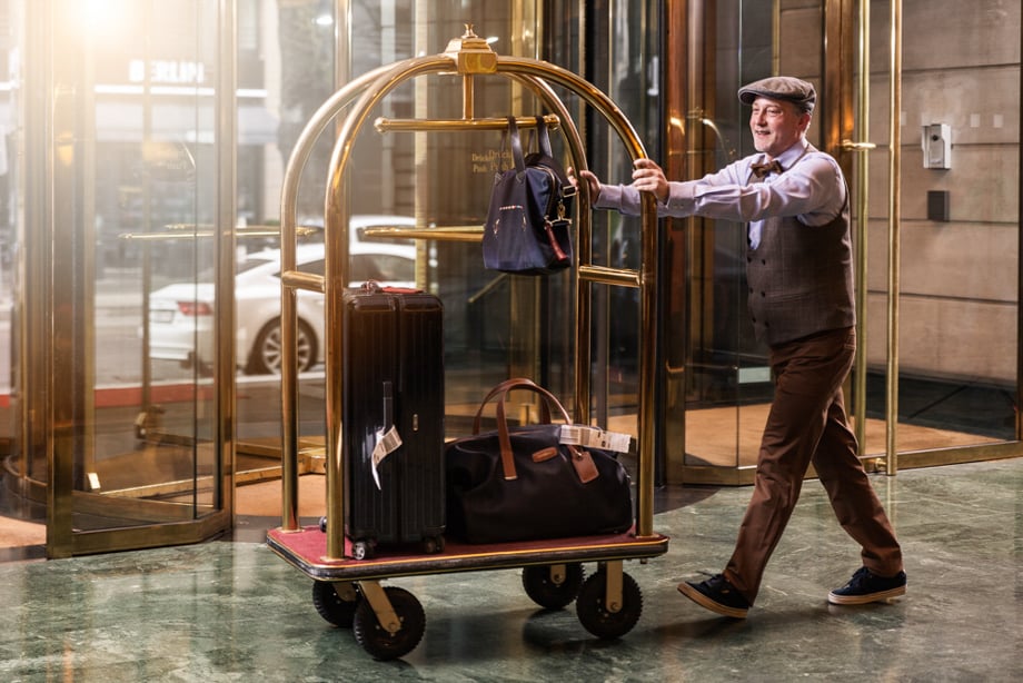 Photographer Aurélien Begot's photo of a bellhop pushing a cart in a hotel lobby with luggage on it. He wears a flat tweed cap, a vest, and a bowtie.