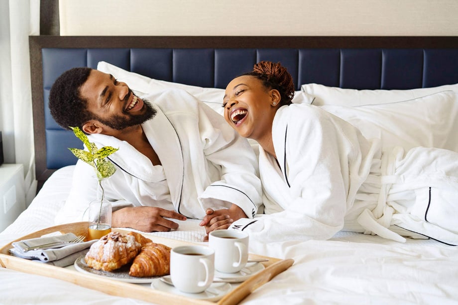 An image by photographer Nicole Loeb of a couple laughing in white robes on a hotel bed. There is a tray with cups of coffee and pastries in front of them and they are holding hands.