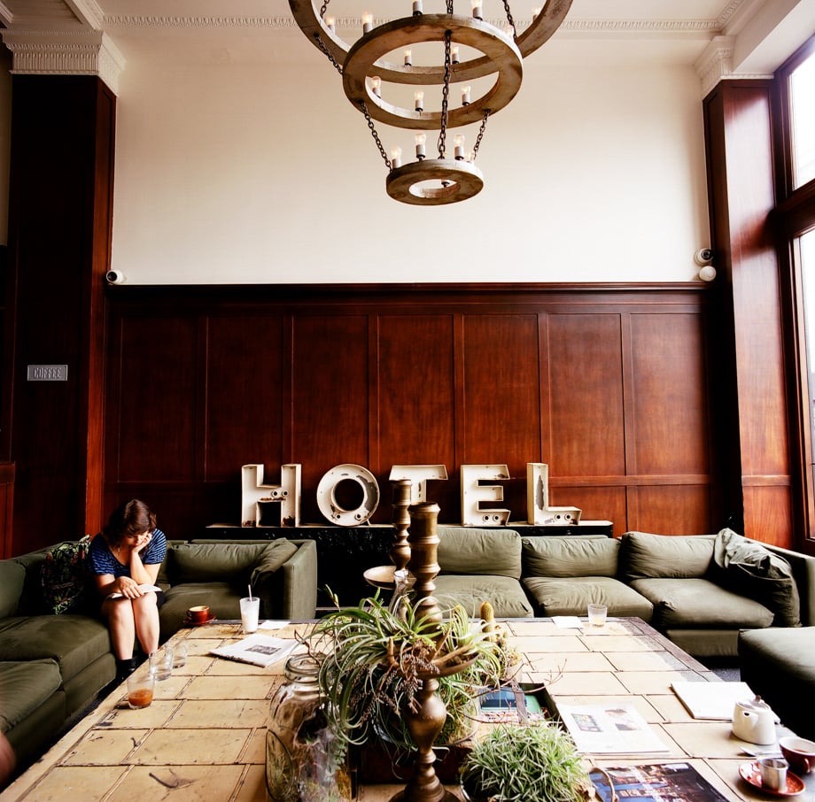 An image shot by photographer Susan Seubert of a common space in a hotel with khaki green sofas, interesting desert-like plants in the center of a large coffee table, and wooden panelling on the walls. On a credenza in the back of the room are large sculptural letters that spell out "HOTEL."