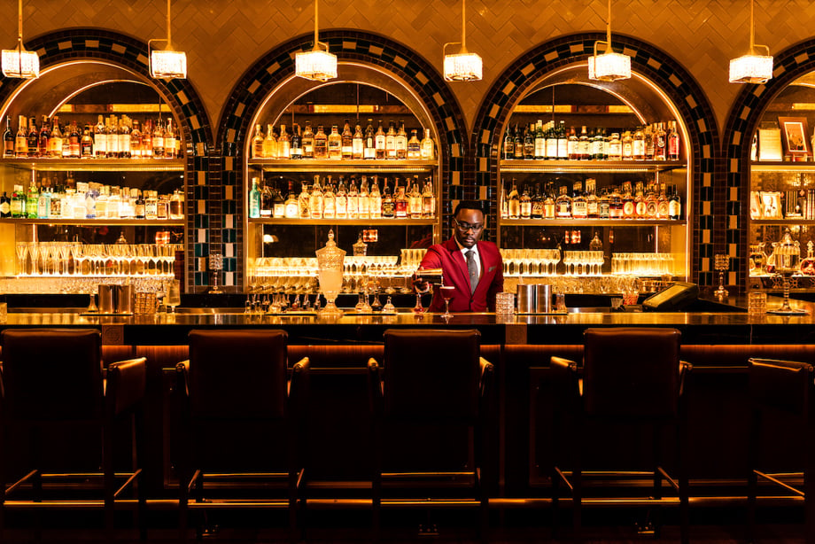 Creative in Place: Be My Guest photographer Ben Weller's photo of a bartender in a vermillion blazer and glasses pouring a cocktail at a warmly lit, stylish bar.