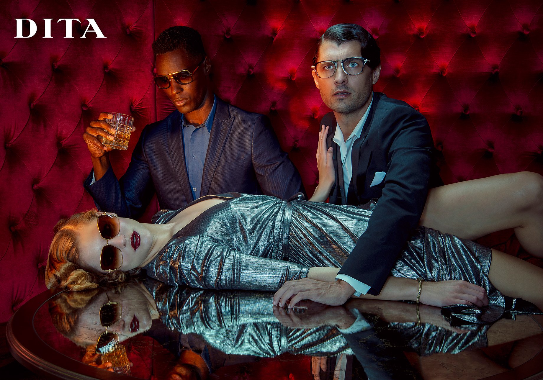 Woman wearing sunglasses, blonde pin curls, and silver cocktail dress lays on table in front of two men in suits and glasses shot by Greg Miles for Dita Eyewear