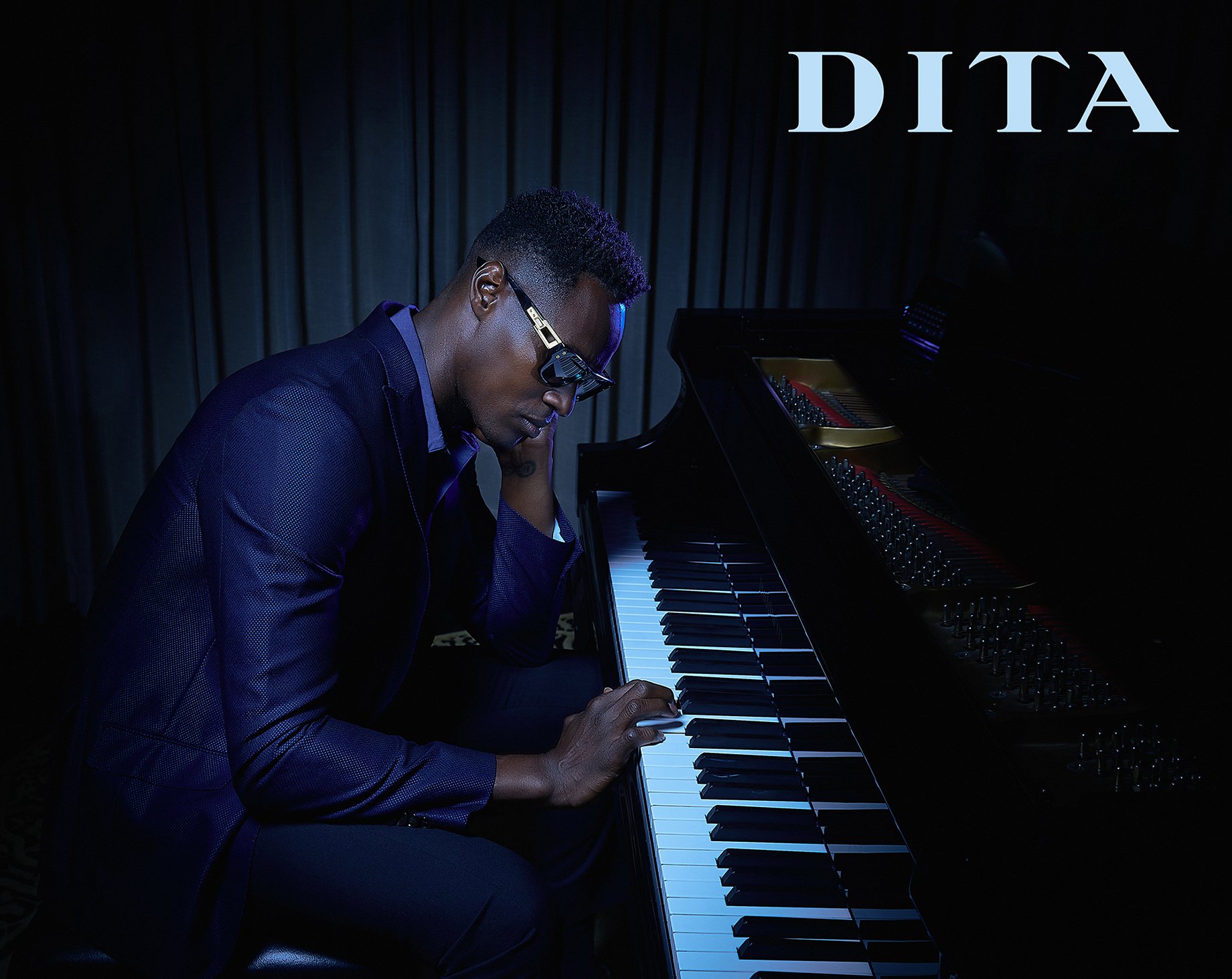 Man in sunglasses and blue suit sits in front of piano with one hand touching the keyboard, shot by Greg Miles for Dita Eyewear