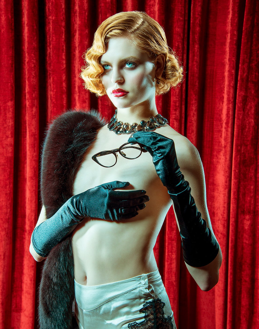 Woman with strawberry blonde pin curls and red lipstick holds cat-eye glasses. She is topless, but is wearing 1920s accessories and covers her chest with one hand clad in long black satin gloves. Shot by Greg Miles for Dita Eyewear