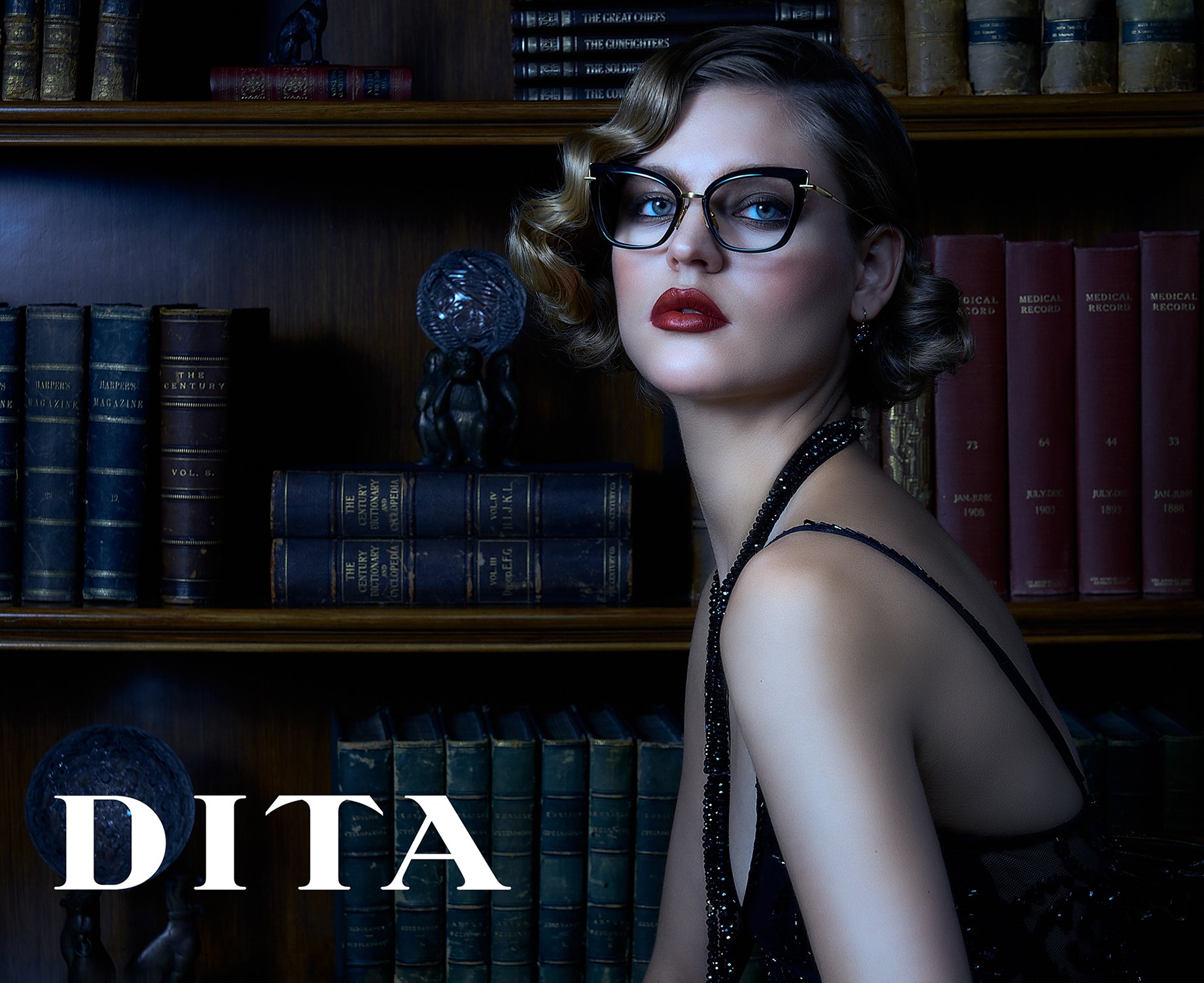 Blonde woman with hair in pin curls wearing glasses, red lipstick, and sparkly beaded black sleeveless cocktail dress sits in front of bookcase full of leather bound books shot by Greg Miles for Dita Eyewear