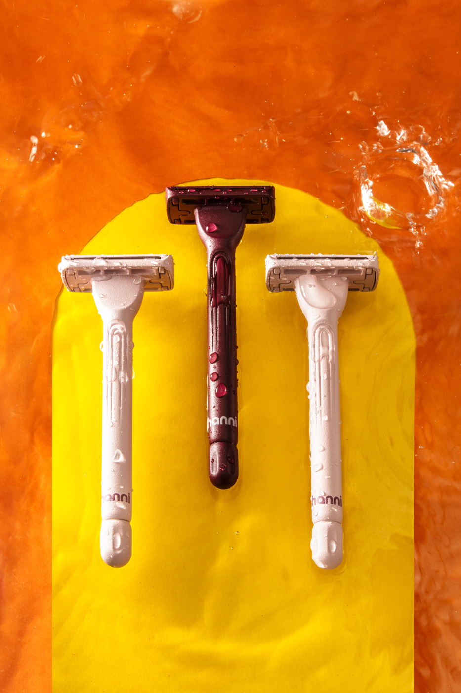 Three Hanni razors submerged in water with splashes on a orange background with a yellow arch shot by Katelin Kinney