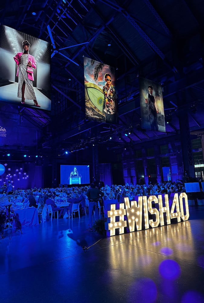 Tear sheet of "wish kid" conceptual portraits displayed at Make-A-Wish 40th Anniversary event space, with seat crowd under dimmed blue lights. 
