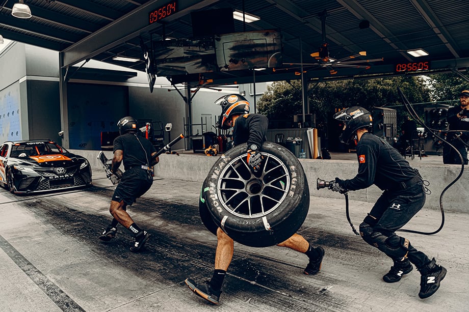 Photographer Will Crooks photographs pit crew members run at oncoming car to practice a pit stop at Joe Gibbs Racing for Road & Track magazine.