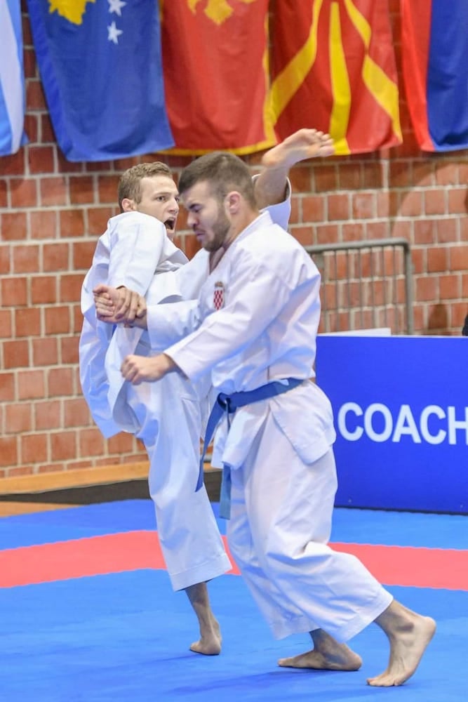Photo of karate champion Mihael Ećimović in uniform high kicking opponent in the head at competition.