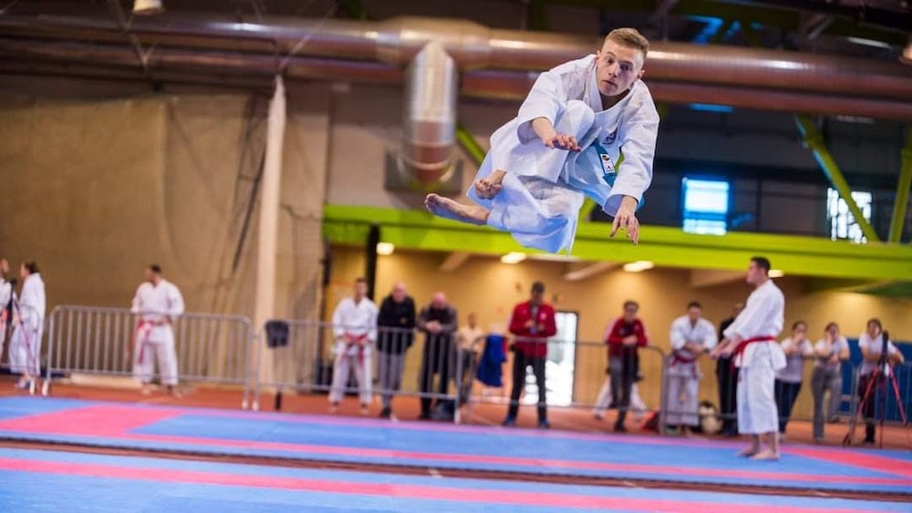 Portrait of karate champion Mihael Ećimović in uniform midair while preforming floor routine at competition.