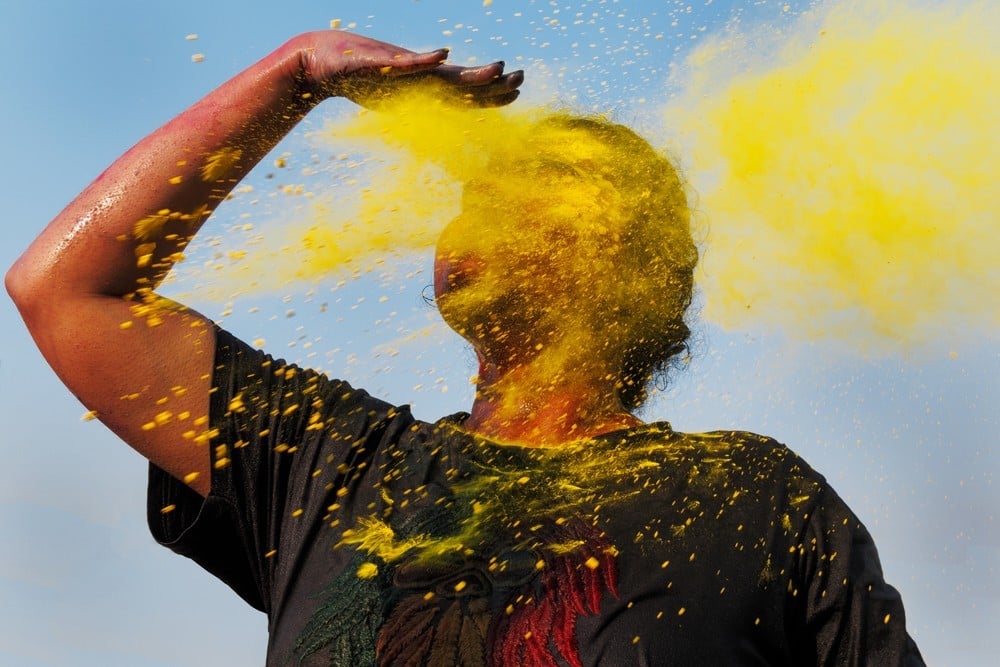 Portrait of figure with hand raised, comically scanning the horizon, as face is met with a cloud of yellow dust, by Mumbai-based portrait photographer Parikshit Rao.
