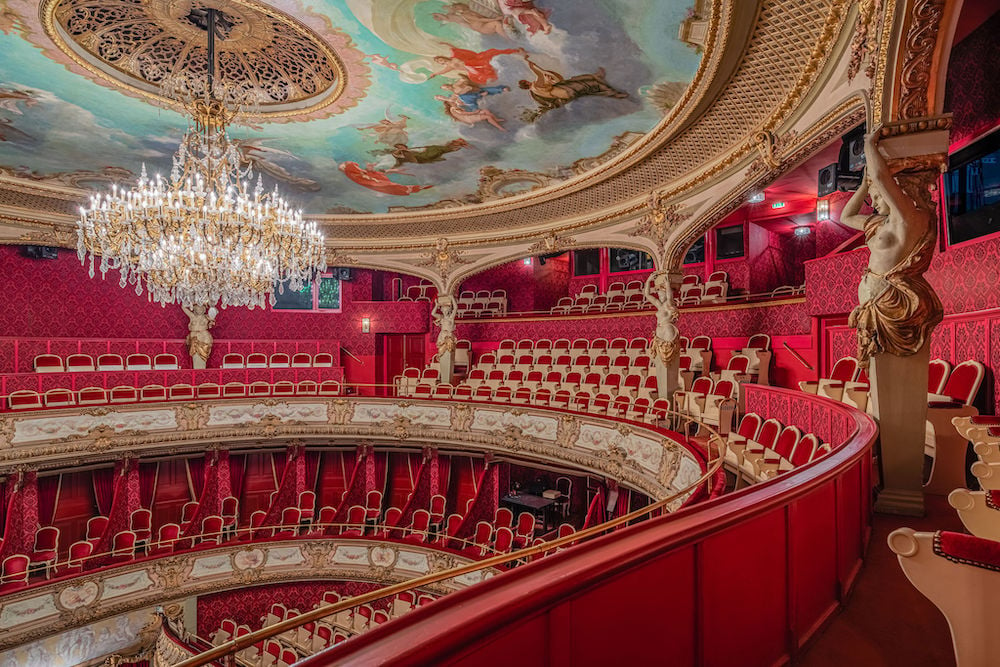Image of picturesque red and gold stadium-style theater interior with crystal chandelier and ornate skyscape ceiling, by Baden-Baden, Germany-based architecture photographer Torben Beeg.