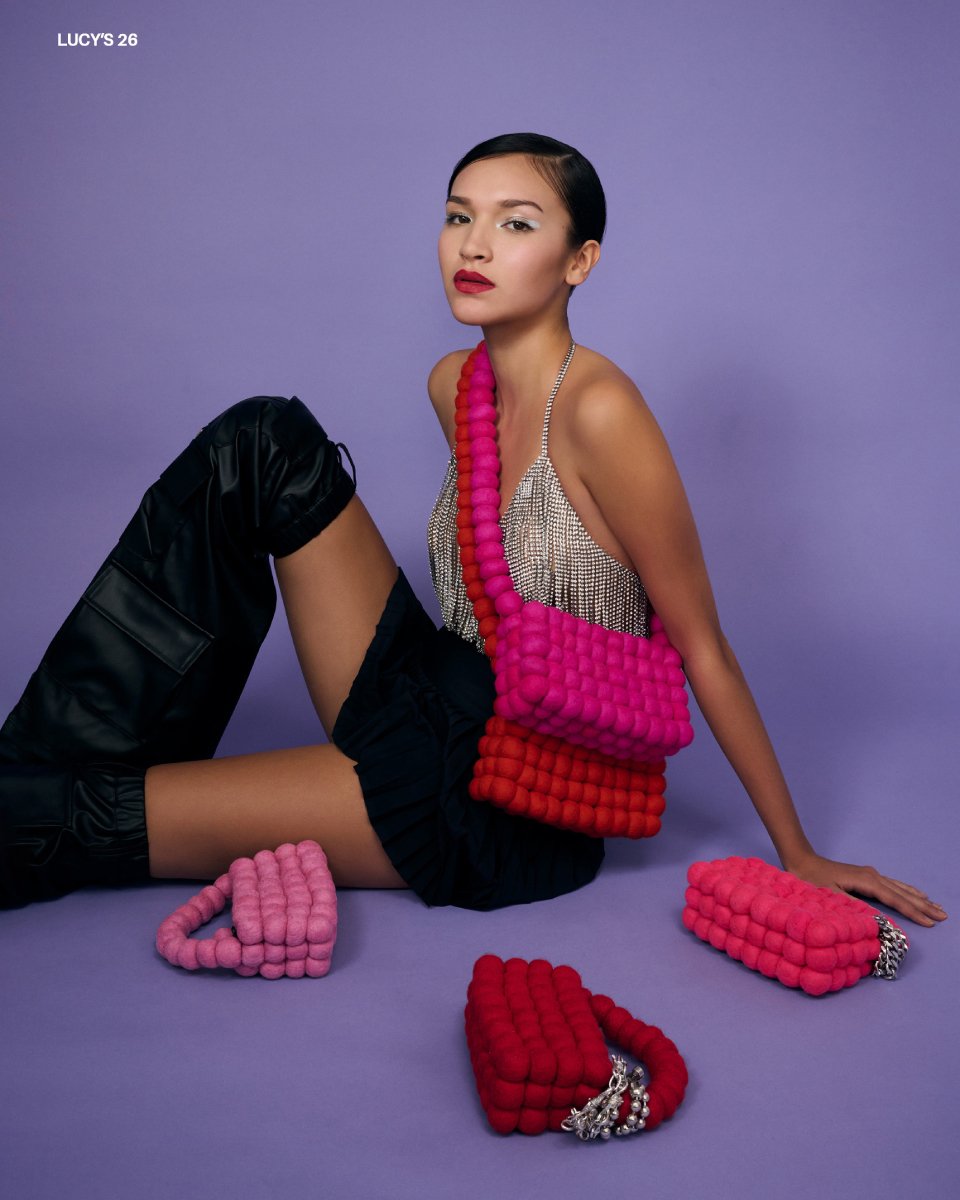 Photo by Matthew Mills of a model wearing two handbags sitting down with three others around her. All are different shades of red.