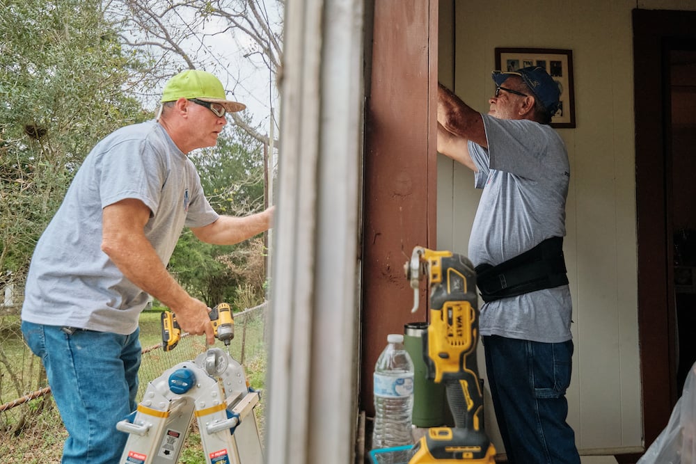 Photo of two volunteers installing a new window, shot from the vantage of a door threshold showing both outdoor and indoor worker, by Mobile, Alabama-based conflict/crisis and humanitarian photographer Dan Anderson. 
