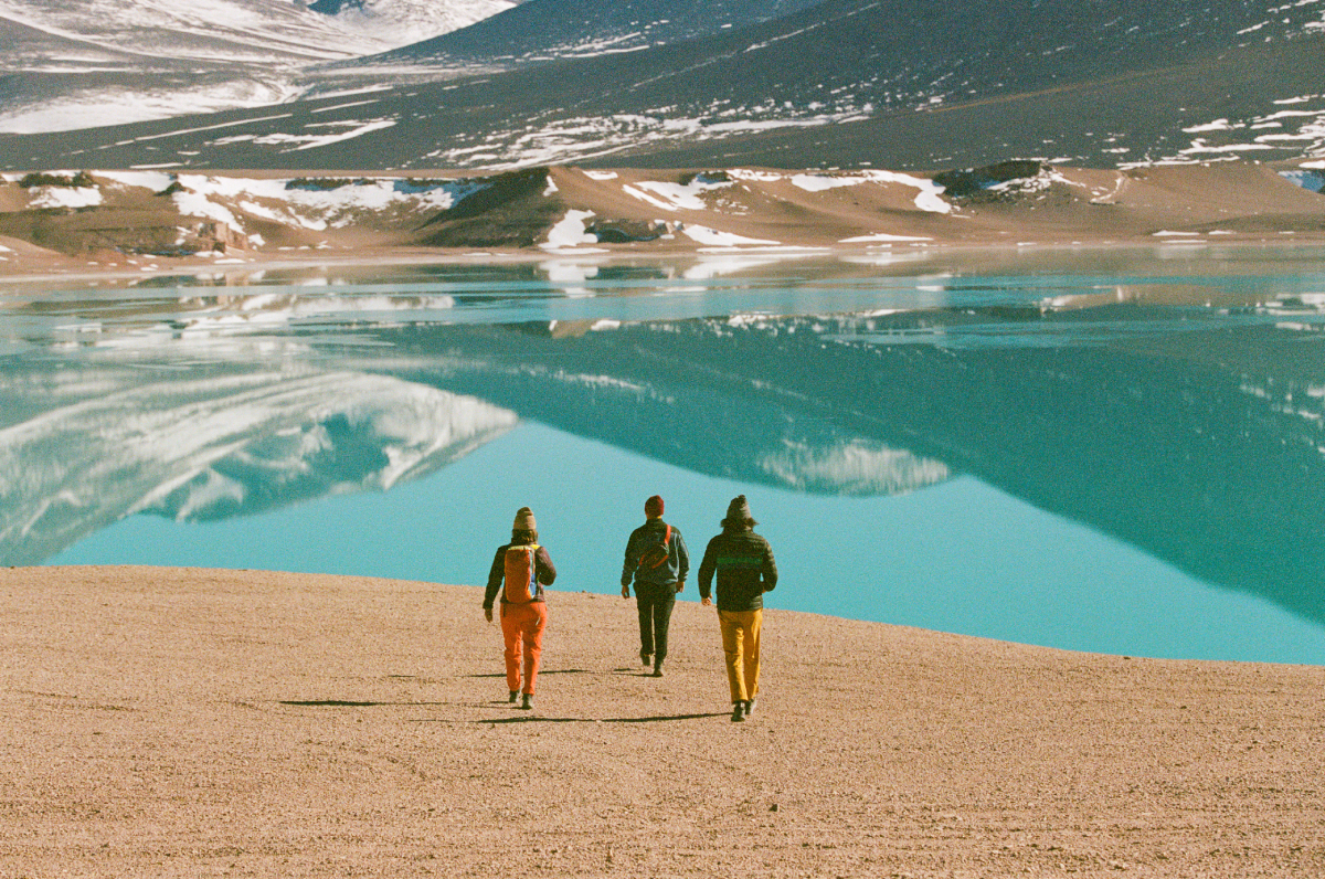 A color photo by Adam Wells of three models walking towards a glacial lake.
